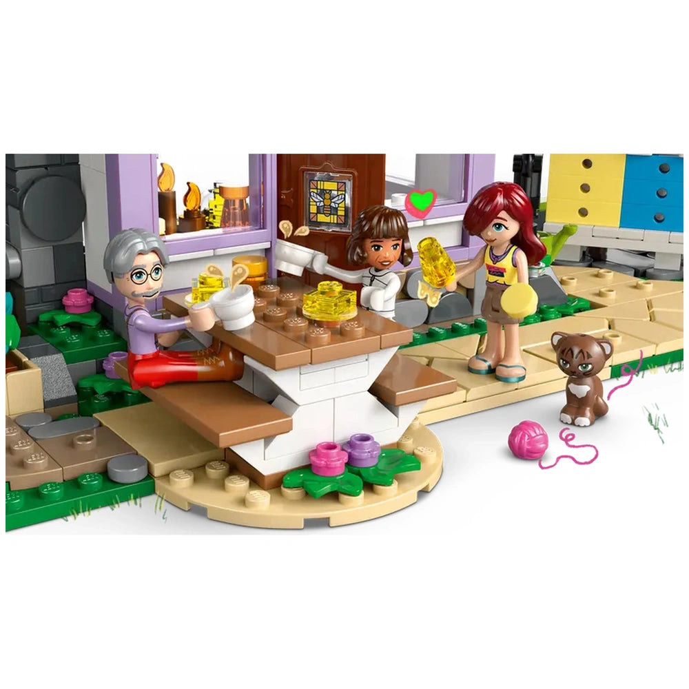 Three LEGO Friends minidolls enjoy a sunny day at a table outside the Beekeepers' House, sharing ice pops. A playful cat sits nearby.