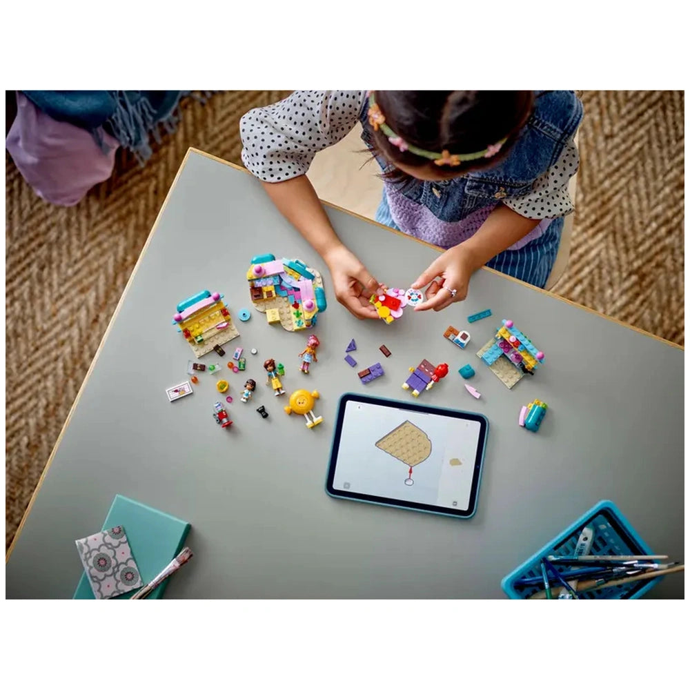 A child with dark hair wearing a polka dot shirt builds with LEGO Friends pieces on a table. Two colorful candy store structures are surrounded by small figures and various accessories. An open tablet displays construction instructions.