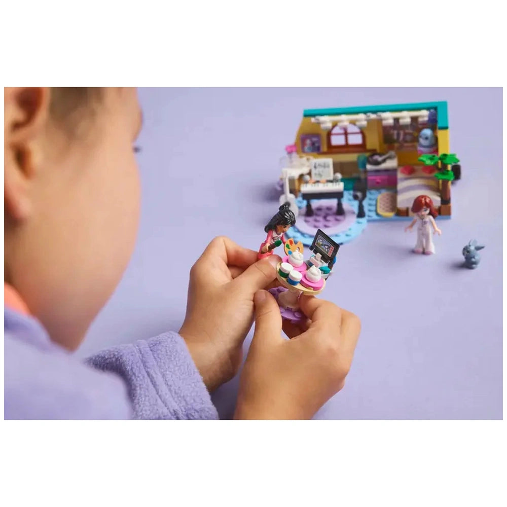 A child in a cozy purple robe holds a LEGO Friends minidoll with a colorful cupcake display, in front of Paisley’s Room set.