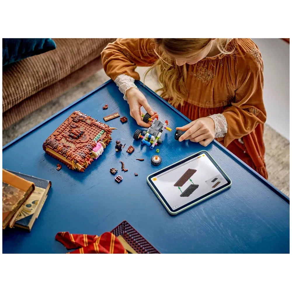 A child in a cozy setting builds the LEGO Harry Potter Chomping Monster Book of Monsters on a blue table, surrounded by scattered colorful bricks. An iPad displaying assembly instructions is nearby, with a partially constructed model featuring bright wheels and detailed pieces. A soft, warm light highlights the creative workspace.