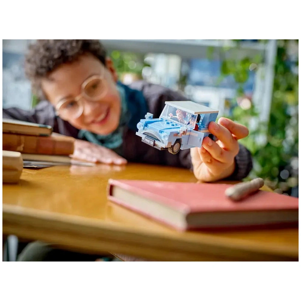 A child with curly hair and glasses smiles while holding a blue LEGO Flying Ford Anglia. The scene features a wooden table with books and a pencil. Green plants are blurred in the background, enhancing the cozy atmosphere.