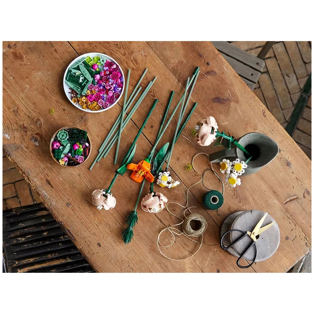 Colorful LEGO flower elements scattered on a wooden table, with completed flower stems and crafting supplies like scissors and twine.