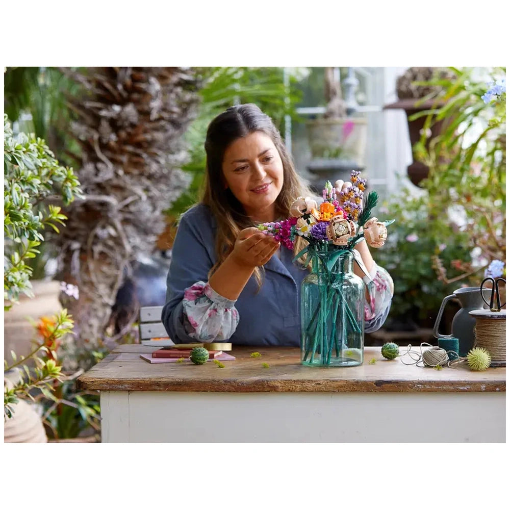 A woman is arranging a colorful LEGO Flower Bouquet in a glass vase on a wooden table surrounded by greenery, showcasing creativity and joy.