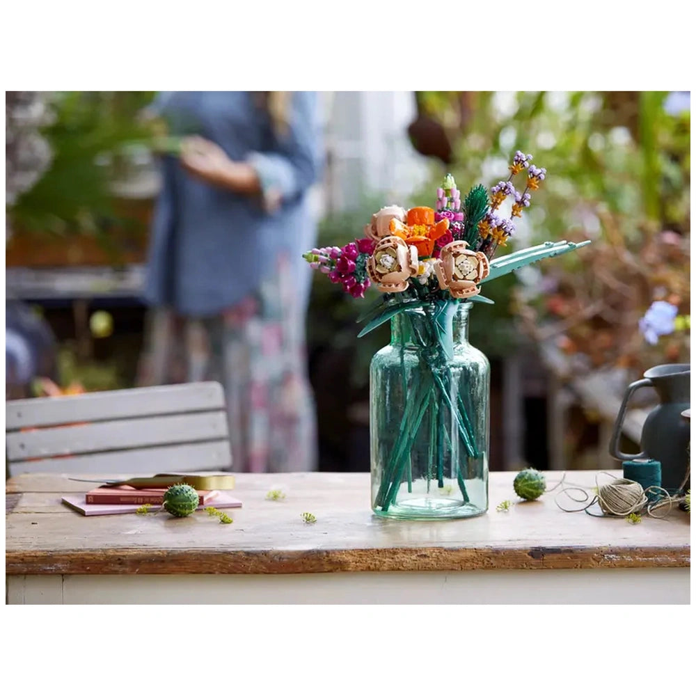 Colorful LEGO Flower Bouquet arranged in a clear glass vase on a wooden table, with craft materials and a blurred background of greenery.