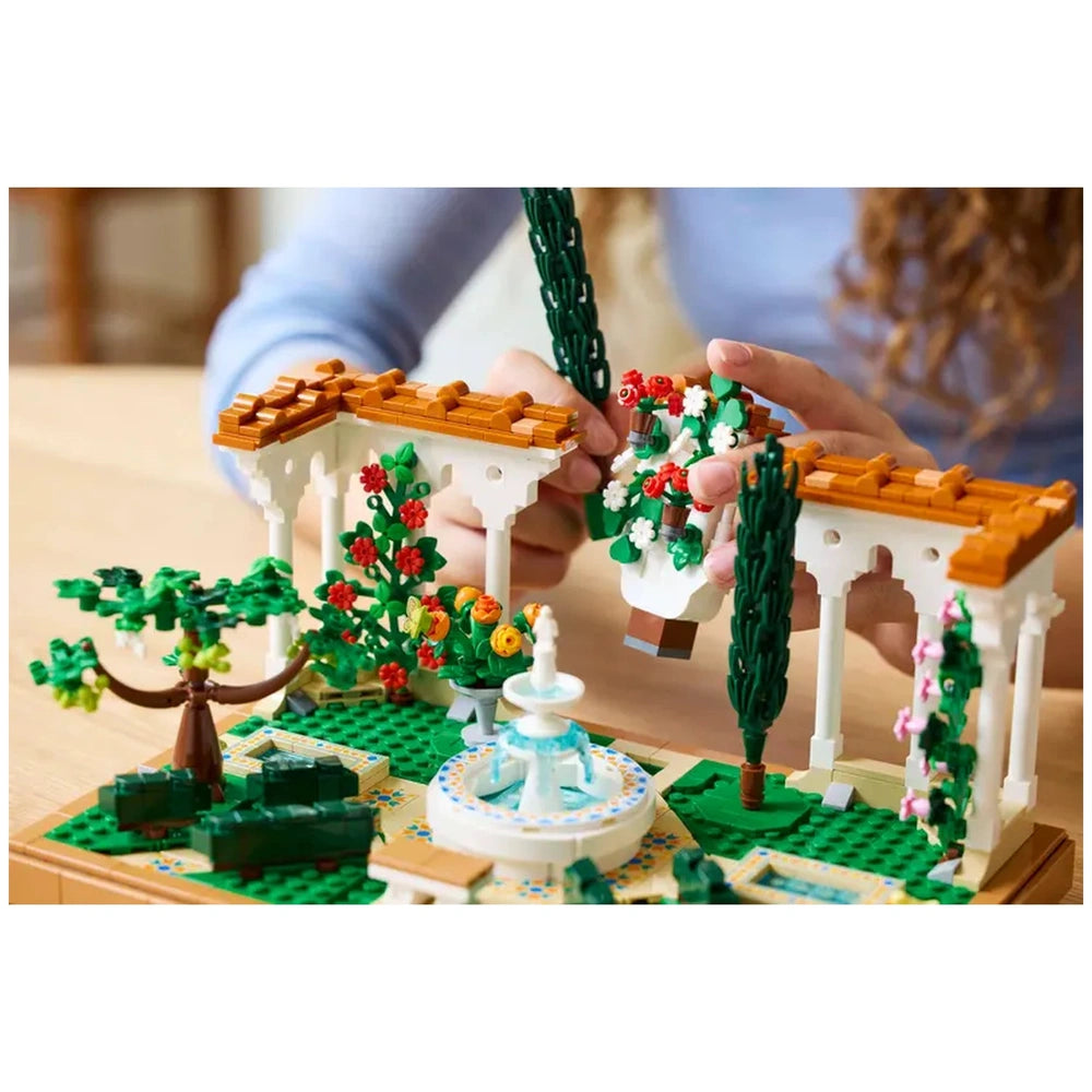 A person with curly hair builds the LEGO® Icons Fountain Garden, showcasing a tiered fountain, decorative plant elements, and colorful flowers in a serene setting.