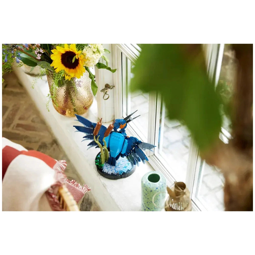 Colorful LEGO Icons Kingfisher Bird construction set perched on a windowsill, surrounded by green reeds and decorative vases.