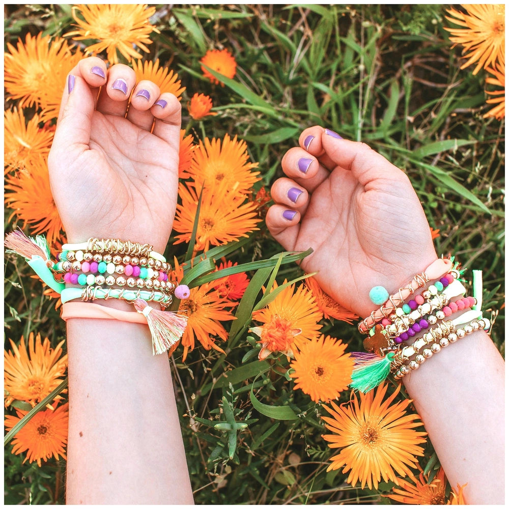 Neo-Brite Chains and Charms showcased with two hands wearing metallic bracelets among orange flowers and green leaves.