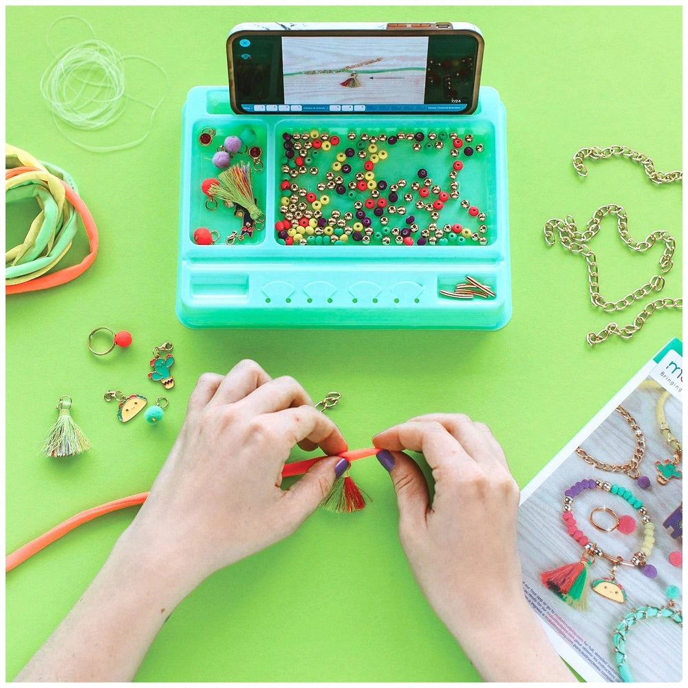 Neo-Brite Chains and Charms in a plastic container on a green surface, with hands holding a red string and bead next to a des