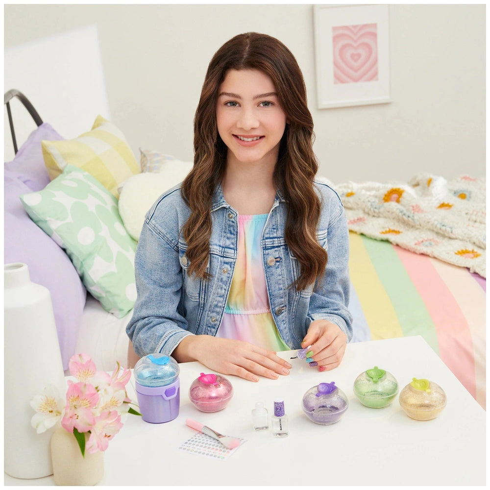 young girl applying MAKE IT REAL Party Nails Glitter Nail Studio products on a white table with colorful nail polish bottles.