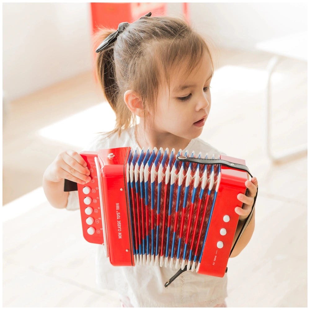 girl plays a red accordion from New Classic Toys, with a music book visible, in a softly blurred room background.