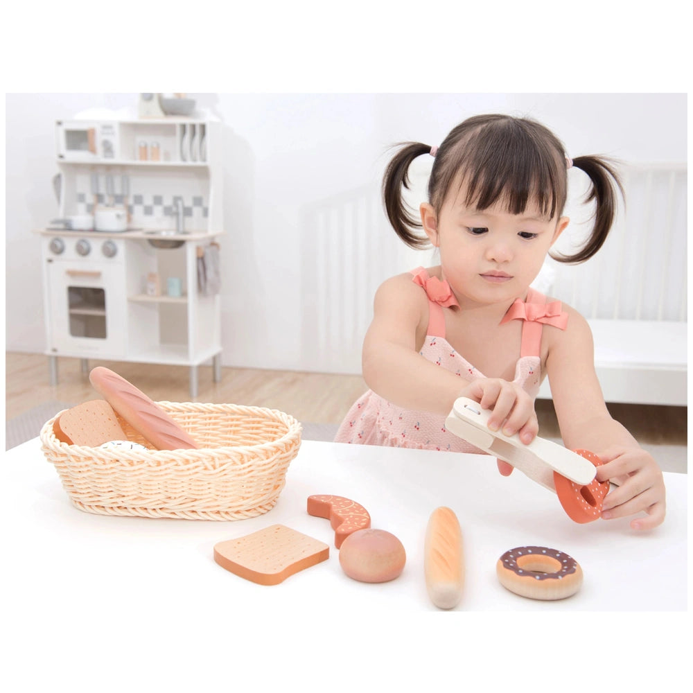 New Classic Toys Bread Basket in a bright kitchen with a girl in pigtails playing pretend cooking at a white table.
