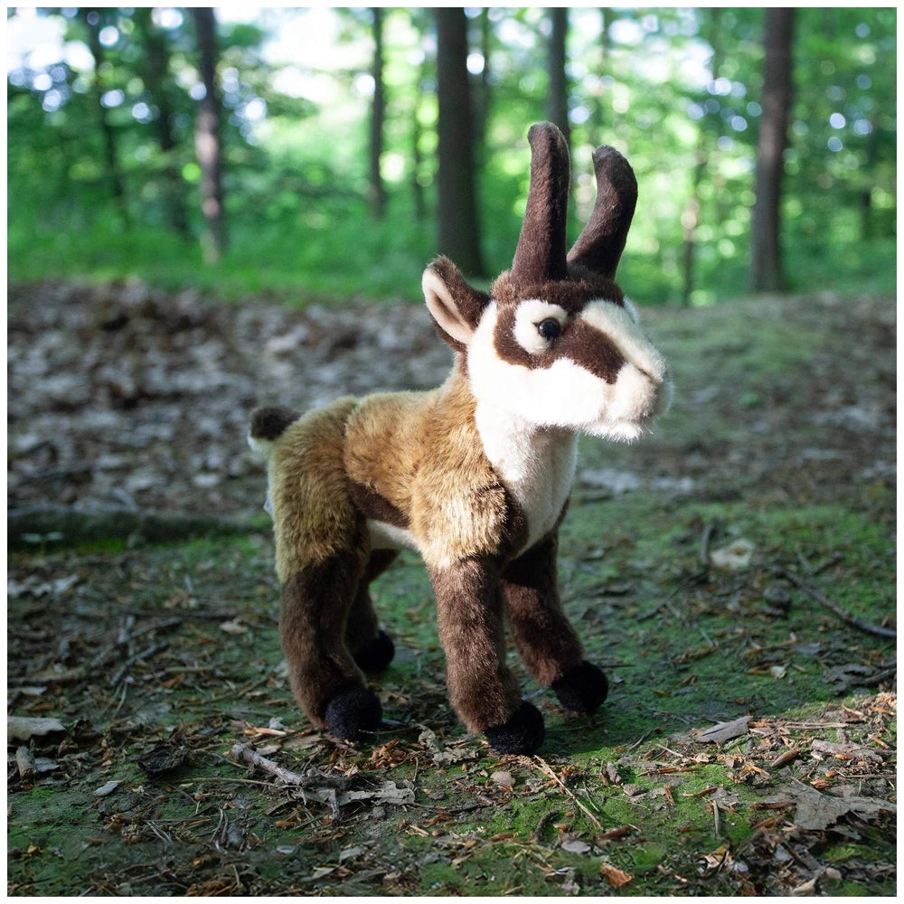 Plush chamois toy with brown and white colors, set on a forest floor surrounded by trees and fallen leaves.