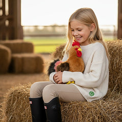 Plush rooster toy held by a smiling girl in a rural setting, surrounded by hay, showcasing its eco-friendly design.