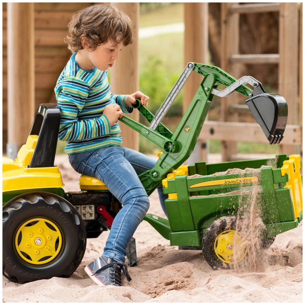 Rollybackhoe John Deere toy tractor with a shovel attachment, occupied by a boy in a striped shirt, on sandy ground.