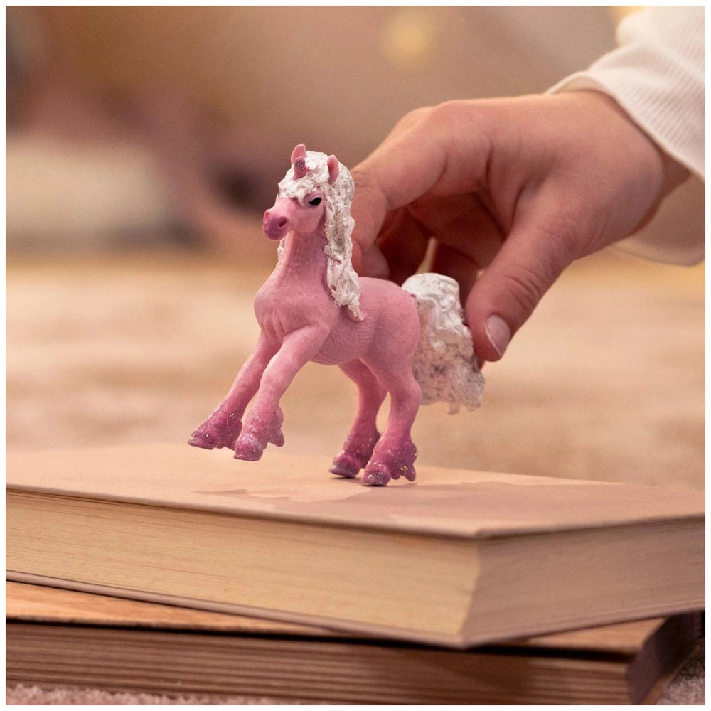 A hand holds a small pink unicorn foal figurine with white accents, set against a softly lit indoor background.