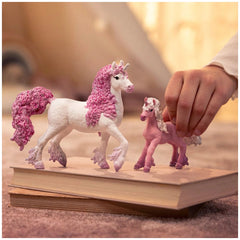 Flower unicorn foal figurine in pink and white held by a hand, resting on a book in a softly lit indoor setting.