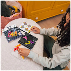 Smart Games IQ Gears puzzle set displayed on a table with a woman assembling colorful crafting materials and an open manual.