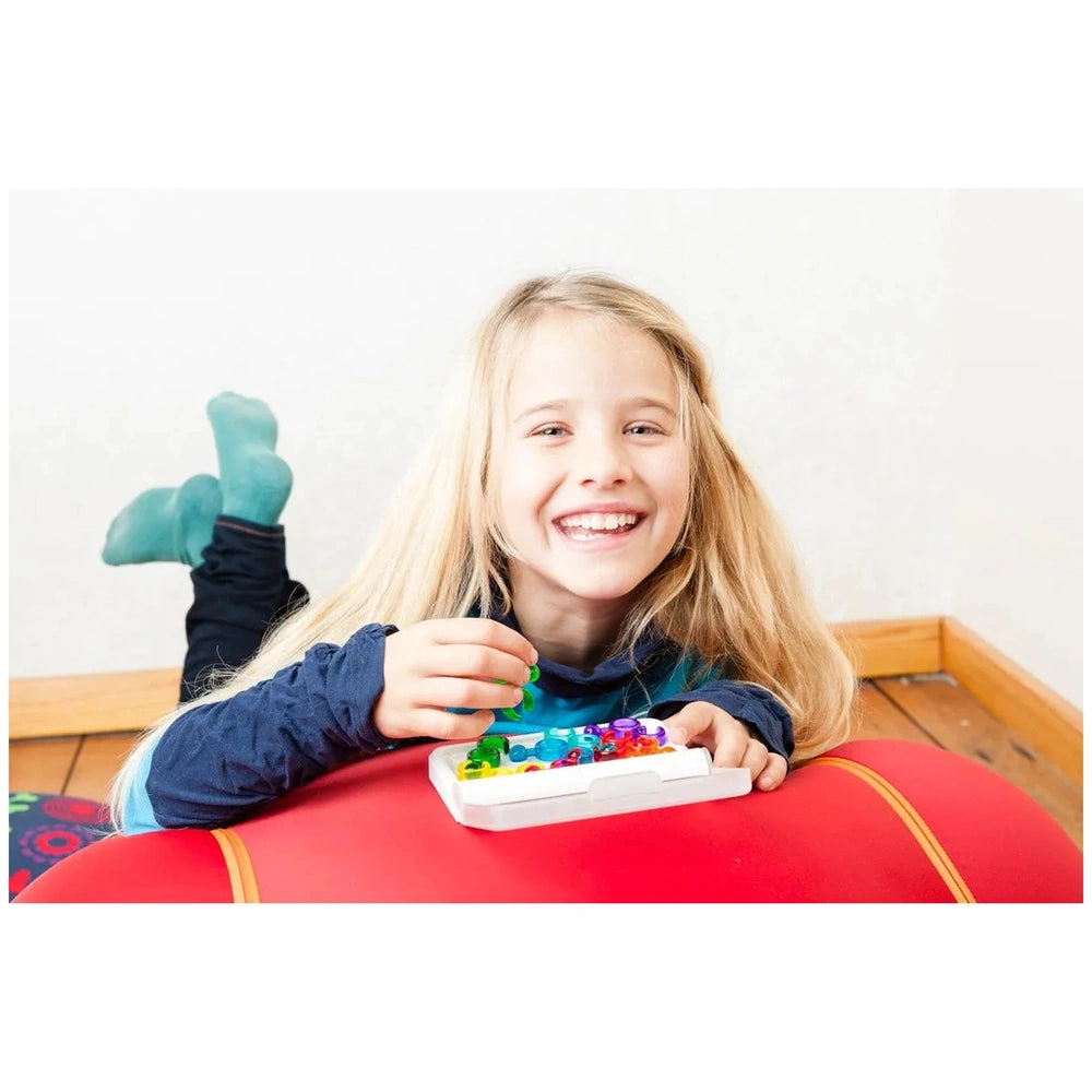 Smart Games IQ Link puzzle is held by a smiling girl in blue socks sitting on a red cushion, enjoying her colorful toy.