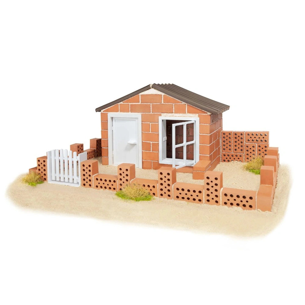 Teifoc beach house model features red brick construction, a brown shingled roof, and is set on a sandy surface with green gra