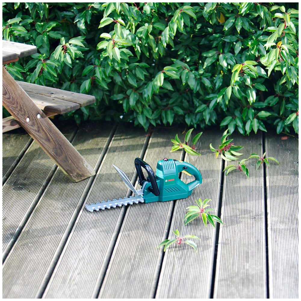 Theo Klein Bosch hedge trimmer on a wooden deck with green leaves, set in a lifestyle scene with an angled view.